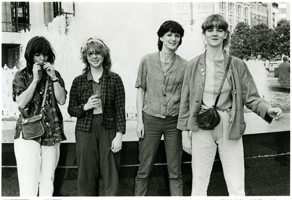 Black and white photo of members of The Raincoats, an all-female artistic post-punk band from late 1970s London. Four members stand (candidly, unposed) in front of a fountain.