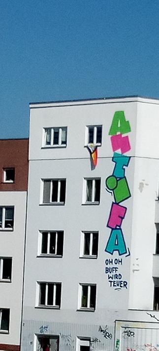 Eine Hausfassade. Von oben nach unten steht in bunten Buchstaben "Antifa". Darunter kleiner in blau oder schwarz außerdem "Oh Oh Buff wird teuer". Eine inklusive Pridefahne hängt aus einem der Fenster.