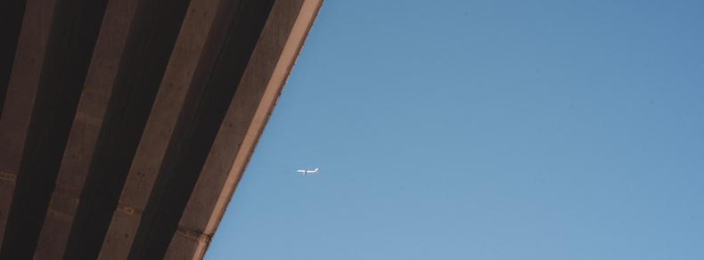 The image shows a clear blue sky with an airplane in the distance. There is a large diagonal structure, possibly an overpass or bridge, on the left side of the image.