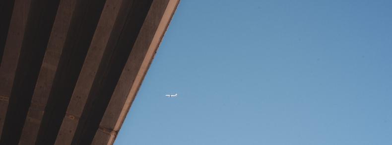 The image shows a clear blue sky with an airplane in the distance. There is a large diagonal structure, possibly an overpass or bridge, on the left side of the image.