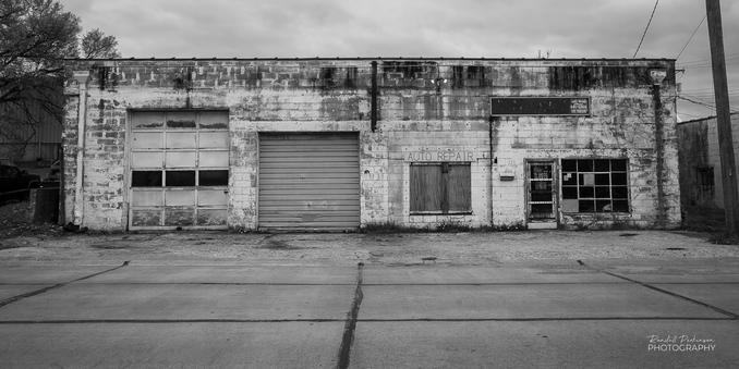 An abandoned auto repair building with two garage doors, two windows, and a door.  The building has a dirty, faded white exterior.