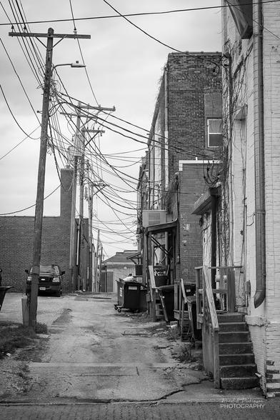 A view looking down an alley behind commercial buildings in a small town.  There are a lot of power lines serving each of the buildings, stairs from back doors, and trash dumpsters.
