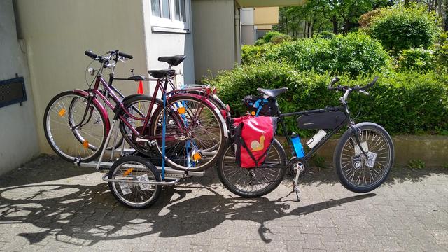 Ein schwarzes Fahrrad mit roten Packtaschen, daran ein Anhänger auf den mit fürs Auto gedachten Dachträgern zwei Fahrräder aufgeladen sind.