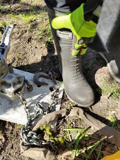 closeup on a black cofra rubber boot with hand wearing a yellow work glove touching the shaft, chain hoist lying on the ground next to the boot