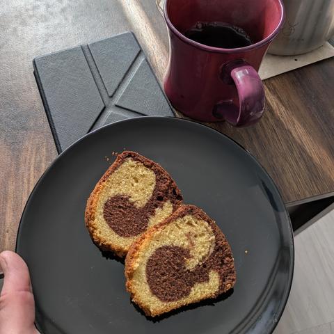 Das Bild zeigt einen kleinen Teller, auf dem sich zwei Stücke Marmorkuchen befinden. Weiterhin einen E-Book-Reader sowie eine Tasse, welche halb mit Tee gefüllt ist. Alles steht auf einem braunen Tisch.
