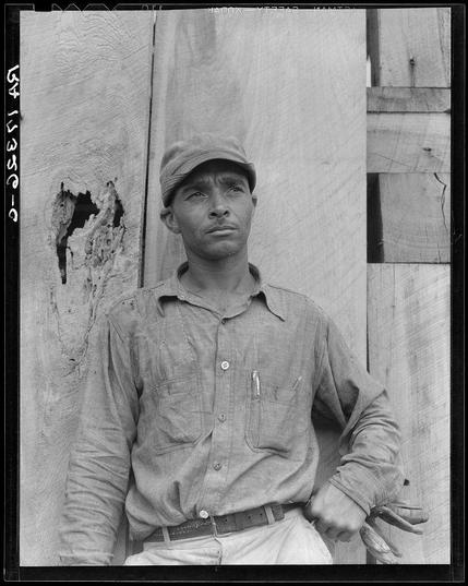 The photograph is a black and white image depicting an older man with weathered skin, dressed in casual work attire. He stands against a rough-textured wall that shows signs of wear, including chipped paint or plaster. His expression appears contemplative as he looks off into the distance over his left shoulder. The lighting casts shadows on his face and emphasizes the texture of his clothing and hands.

The man wears a cap with a worn look, suggesting outdoor labor use. His shirt is buttoned up to the collar but has visible wear marks indicating frequent handling or physical activity. He clasps one hand around his belt while resting the other arm across his chest, adding to the casual yet reflective pose.

The photograph exudes an aura of rural life and hard work, potentially highlighting themes from a historical perspective on American agricultural workers during the early 20th century. The reference indicates that this individual was likely part of a cooperative farm in Hillhouse, Mississippi, which may provide context for his attire, demeanor, and environment depicted.

There is handwritten text at the top left corner: "649538-CO-1", possibly indicating an archival or cataloging code related to Dorothea Lange's photography work.