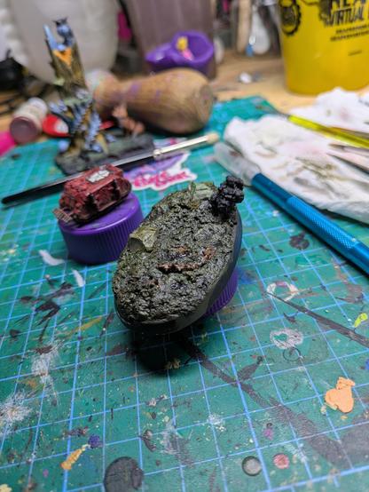 A partially painted oval base with mud, rocks, and Tufts. It has an unpainted explosion plume and a tiny red tank is visible behind slightly out of focus.