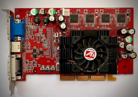 Es wird eine AGP Grafikkarte der Firma ATI aus dem Jahr 2003 gezeigt. Diese hat eine rote Leiterplatte, einem Lüfter, verschiedenen elektronischen Komponenten, Anschlüssen für die Videoausgabe und den typischen dreiteilen AGP Stecker an der unteren Kante.