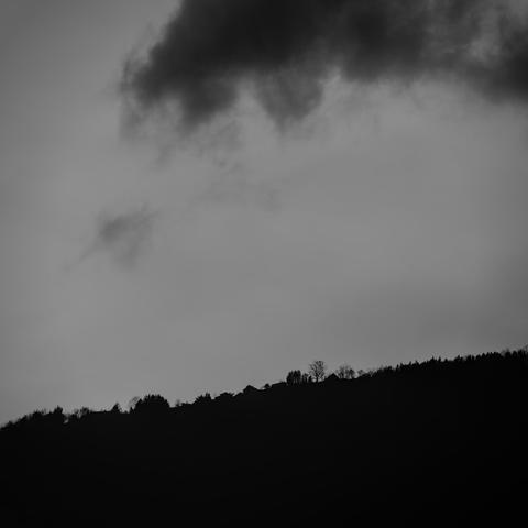 Photo noir et blanc d'une crête en ombre chinoise. Quelques arbres isolés et des toits se découpent.
Dans le ciel autrement bleu, un unique nuage se délite, sombre.