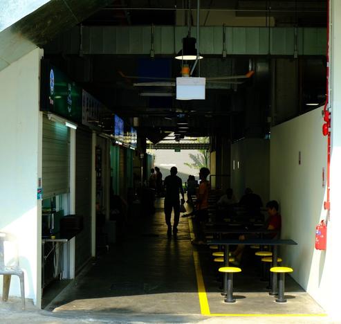 Taken during the bright sunlight of mid-afternoon, looking into the dark interior of a hawker center. The silhouetted figure of an old man is walking away from the camera.