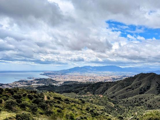landscape from above a hill showing a coastal city and some mountains in the background.