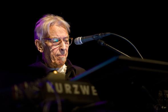 Der Musiker John Cale mit Brille und weißen Haaren. Nahaufnahme vom Gesicht. Er sitzt hinter einem Keyboard, vor sich ein Mikrofon. Der Rest des Bildes ist in Dunkel getaucht.