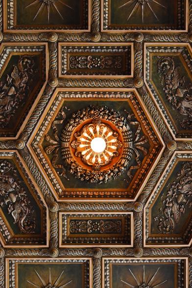 Color photo looking directly up at a profusely decorated plasterwork ceiling which features a glowing light fixture - with individual bulbs radiating out from its center - set into a hexagonal coffer. All of the ceiling’s raised, Neoclassical design elements have been gilded.