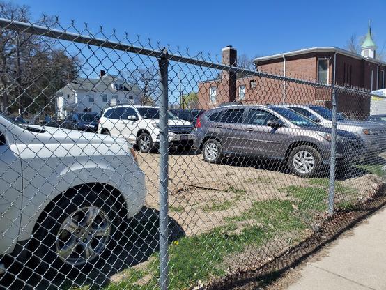 The yard behind a brick a brick church, enclosed by a chain link fence. Cars are parked throughout the yard.