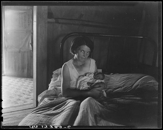 The image depicts a woman seated on a bed in what appears to be an interior room, possibly within a modest home or cabin. She is holding and gently looking down at a small infant wrapped in cloth. The woman has short hair styled closely to her head, wears minimal makeup with attention given to eye shadow and lipstick, and dons a sleeveless white top that exposes the upper portion of her arms.

Behind her on the wall hangs an empty bag labeled "Corduroy," suggesting she may be involved in some form of manual labor or has recently been shopping. The room is dimly lit with natural light coming through what seems to be a doorway, revealing a glimpse of another room or outdoor space beyond it. There's visible wear on the bed linens and wooden furniture around her, indicating modest living conditions.

The photograph carries historical weight as evidenced by its quality, black-and-white nature, and textual information at the bottom which reads "1520 3/6" in what could be a reference to an inventory or cataloging system.
