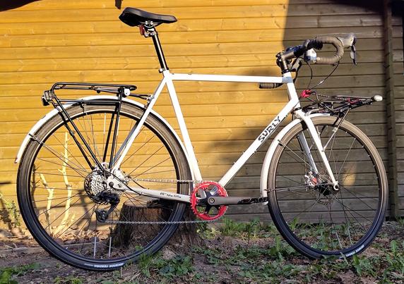 Ein weißer All-Road-Bike-Rahmen mit roten Details an Kettenblatt, Flaschenhalterschräubchen, Spanngurtkarabinern, Smartphonehalterung und Bremseinstellungsrädchen. Abgelichtet im Garten vor einer hellen Holzgartenlaube