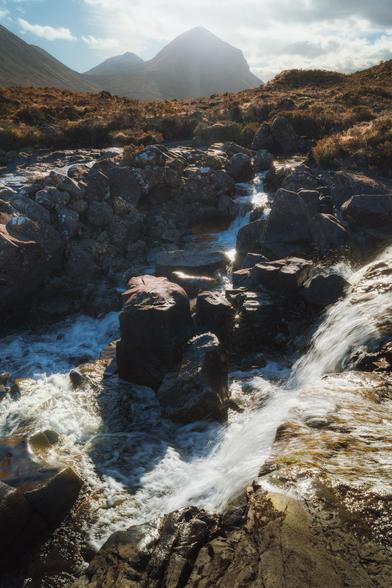 This image, taken at Sligachan on the Isle of Skye, Scotland, captures a lively stream tumbling over dark, rugged rocks, creating a series of small, frothy waterfalls in the foreground. The sunlight glints off the wet stones and water, adding a sparkle to the scene. In the background, the rounded, softer shapes of the Red Cuillins rise against a partly cloudy sky, their peaks bathed in a soft, hazy light that contrasts beautifully with the sharp textures of the moorland and rocks below.