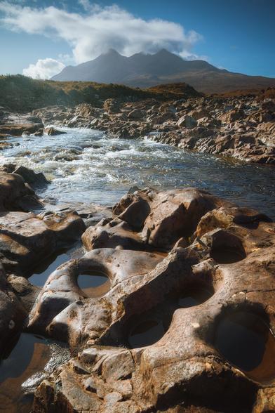 This image, taken at Sligachan on the Isle of Skye, Scotland, looks towards the brooding Black Cuillins, whose sharp peaks are partly veiled by swirling clouds beneath a vivid blue sky. In the foreground, weathered reddish rocks are dotted with smooth, water-filled potholes, leading the eye towards a gently flowing river sparkling in the sunlight. The scene is bathed in a warm, crisp light that highlights the rugged textures of the rocks and the golden-brown moorland stretching towards the distant mountains.