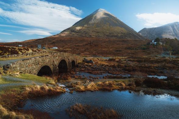 This image, taken at Sligachan on the Isle of Skye, Scotland, shows a charming old stone bridge with three arches spanning a shallow, rocky river under a brilliant blue sky. In the background, the broad, rounded slopes of the Red Cuillins rise steeply, their rugged surface catching the light and partially dusted with snow. The surrounding landscape is a tapestry of golden-brown moorland and patches of rough grass, while a few vehicles and a white building are visible near the roadside, hinting at the area's popularity with visitors.