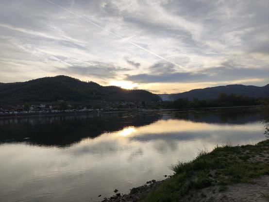Ein großer Fluss, die hinter einem Hügel untergehende Sonne spiegelt sich im Wasser.