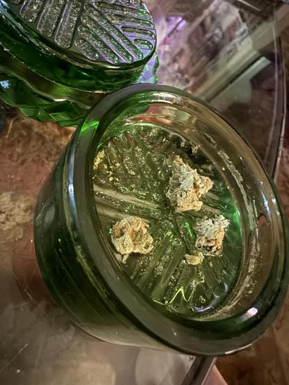 A round and bottle-green glass canister with ridges on the inside, there are three small buds of cannabis sitting in the grinder. The top sits with it on the clear glass table, above and to the left.