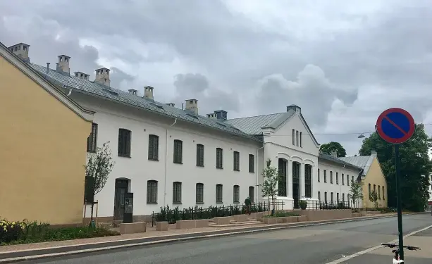 The image depicts a building with white walls and a sloped roof, characterized by several chimney structures. It's set beside a road with a red no-parking sign, and there are small plants and greenery along the sidewalk. The sky appears to be overcast with gray clouds. The architecture features large windows, and there are other structures visible in the background.

Image Credits: Wikimedia / Wolfmann / CC BY-SA 4.0