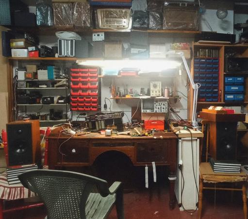 Fotografía en la que aparece una mesa de madera antigua con un montón de aparatos, herramientas y objetos de electrónica. En las paredes también hay cajas que guardan piezas y estanterías con radios y aparatos electrónicos antiguos, algunos envueltos en plástico. También hay dos altavoces a los lados de la mesa, situados en sillas viejas y apoyados en libros antiguos, y una silla de jardín de plástico verde y envejecida. Toda la imagen está atestada de objetos y de cosas generando un "horror vacui".