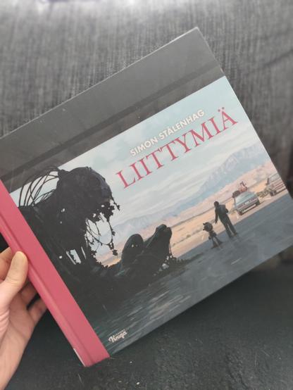 Simon Stålenhagin Liittymiä-kirja Katjan kädessä. Kirja on kooltaan iso: leveydeltään normaalia suurempi, kuin kuvakirja. Kannessa on nuori ihmishahmo ja pienempi robotti, jotka ihmettelevät jättikokoista, istuvaa robotin kaltaista olentoa, joka näyttää sammuneen. Taustalla kuivan näköinen maisema ja auto.