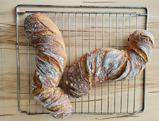 Zwei schlanke, in sich gedrehte Brotlaibe, Ciabatta ähnlich. Sie haben eine leichte S-Form und liegen auf einem Gitterrost auf einer Holzfläche.