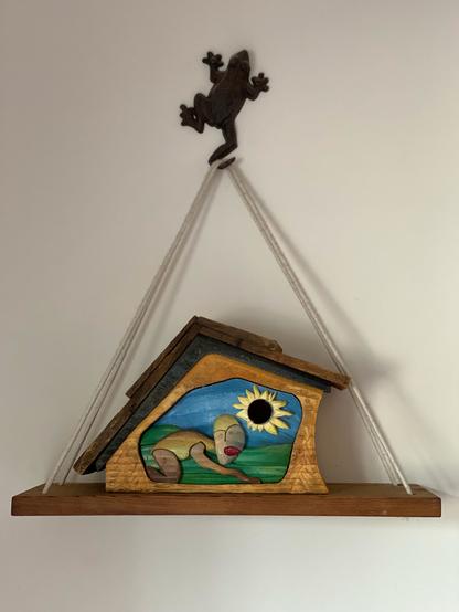 A wooden bird house/art work showing a stylized human figure bowing before a sun that looks like a sunflower. The background is in undulating shades of green below with a bright blue sky. The bird house has an asymettrical shape and a wooden shake roof. It sits on a wooden shelf suspended by ropes from the foot of a metal frog.
