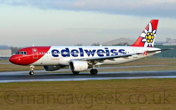 Side view of a white, twin engined jet airliner moving at high speed from left to right on a black runway.
The plane has a red forward fuselage, with a white flower with a yellow middle on the forward door.
There are blue, billboard-style "Edelweiss" titles on the fuselage, while the tail is red, with a larger, white flower with a yellow middle.
The vertical endplates on the wingtips have a smaller version of the same red background and white and yellow flower.
The registration "HB-IHY" is visible on the lower rear fuselage, just aft of the rear door, next to a small Swiss flag.
Green grass fills the foreground, with more grass and taxiways in the background, leading off to grey buildings in the distance, under grey skies.