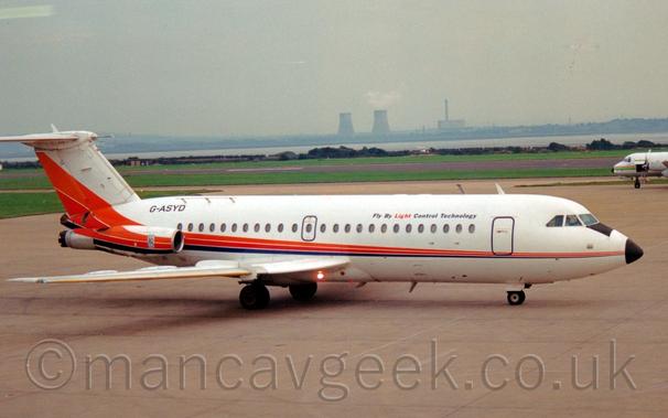 Side view of a white, twin engined jet airliner with the engines mounted on the sides of the rear fuselage, taxiing from left to right.
The plane is mostly white, with an orange, red, and blue stripe running along the body, slowly getting wider as it goes along, eventually sweeping up into the lower reaches of the tail, as well as running along the engine pods.
There are black "Fly By Light Control Technology" titles on the upper forward fuselage, with the word "Light" in red.
The registration "G-ASYD" is on the upper rear fuselage, in black.
Grey concrete apron fills the foreground, with large areas of green grass and a black runway in the background, leading up to a river in the distance, with an industrial landscape in the distance, under grey skies.