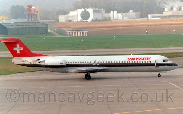 Side view of a white, twin engined jet airliner with the engines mounted on the sides of the rear fuselage, taxiing from left to right.
The plane is mostly white, with a grey belly and brown and black stripe running along the body and the lower part of the engine pods.
There are red "SwissAir" titles on the upper forward fuselage, and black registration "HB-IVC" on the rear fuselage.
There are also small "Fokker 100" titles on the forward part of the engine pods.
The tail is red, with a large white cross in the middle.
A large expanse of concrete apron fills the foreground, with green grass and a runway in the distance leading up to a crop field in the distance.
Right in the background is a very distinctive white building, with a black glazed circle in the middle of a square block.