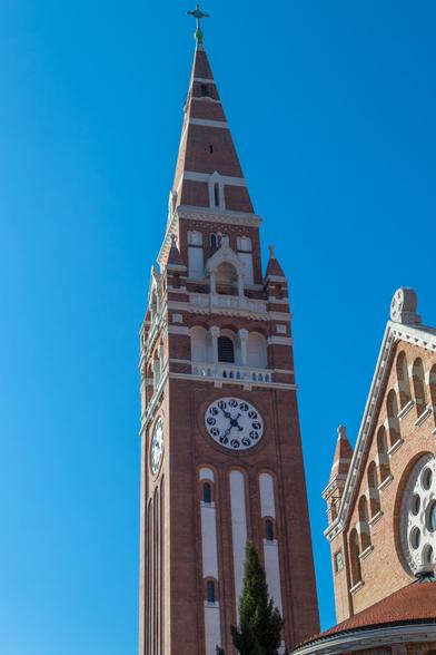 Brick clock tower of Votive Church and Cathedral of Our Lady of Hungary with cross on top under a clear blue sky, showing 10:25 AM.
