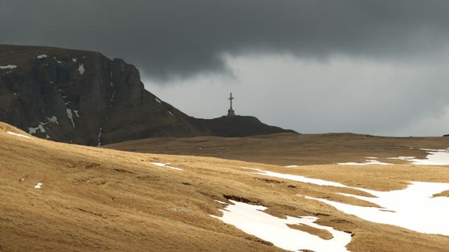 Blick über eine noch gelbe Hochgebirgswiese, teilweise mit Schnee bedeckt, zu einem Felsmassive, auf dem ein Gipfelkreuz steht. Dunkle Wolken am Himmel.