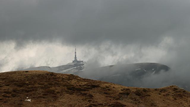 Blick über eine braune Hochgebirgswiese zu einem Felsmassive, welches in den grauen Wolken verschwindet, zu einem Funkturm.
