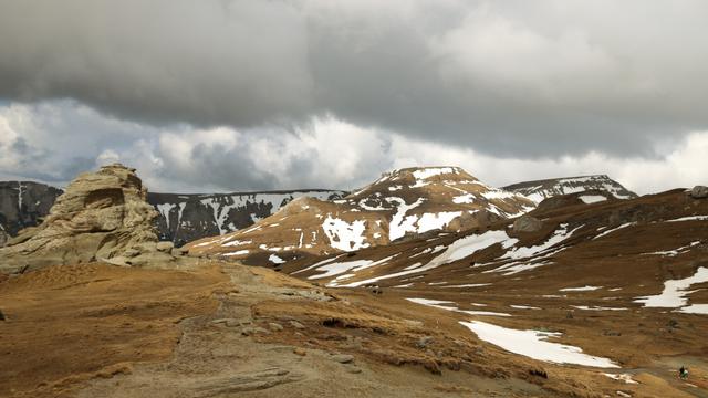 Hochgebirgswiese gelb und braun mit Schneeresten. Felsformationen und Bergkuppen.
