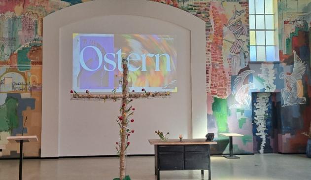 Ein Kreuz aus zwei dicken Ästen, mit Maschendraht umwickelt, Tulpen in verschiedenen Farben hineingesteckt. Rechts davon eine Werkbank mit Schraubstock und Kerze. Im Hintergrund diesmal ein buntes Bild mit dem Wort "Ostern". Am der Wand ein Wandgemälde mit Szenen aus Berlin und einer einstürzenden Mauer und einem Engel