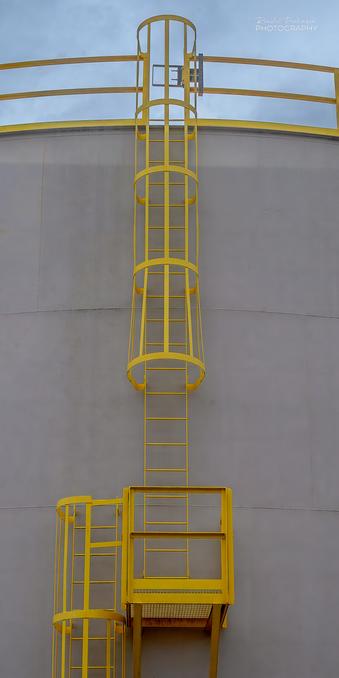 A yellow ladder with a mid-height platform and safety cages rises to the top of an oil storage tank.