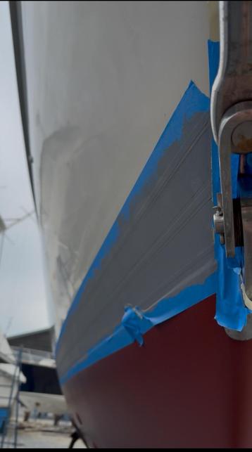 Close-up view of a boat's hull with blue painter's tape applied along the area where a stripe would go. The stripe is painted gray, indicating preparation for painting or maintenance.
