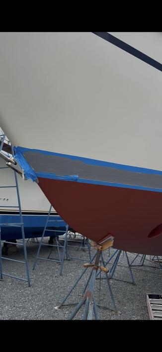 A boat hull is partially painted, with blue tape applied along the area right above the waterline where a stripe might go. The red bottom of the hull is visible, and the boat is supported by metal scaffolding on a gravel surface. Another boat can be seen in the background.