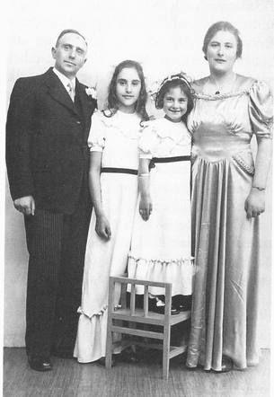 Four people standing in a line. A man in a suit, two girls in white dresses (the second is standing on a small chair), and a woman in a long evening dress.