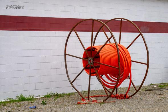 A metal spool containing a small amount of orange fiber optic conduit sits on a gravel lot next to a white concrete block building with a red stripe running horizontally across it.