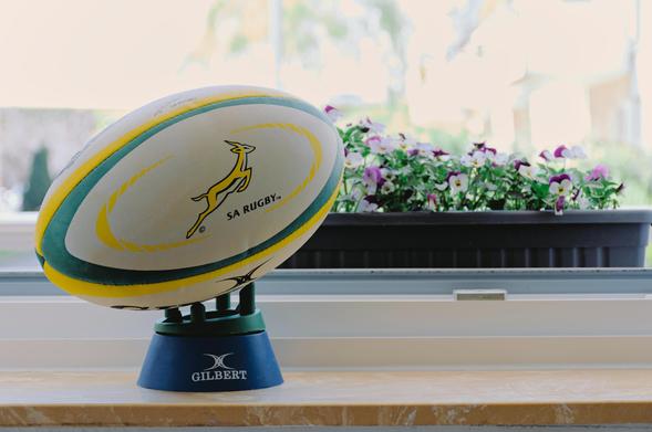 The image shows a rugby ball placed on a stand. The ball has a logo with a springbok and the text "SA RUGBY" on it. The stand has the brand "Gilbert" on it. In the background, there is a planter with purple and white flowers.