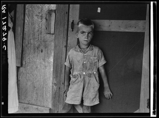 This black and white photograph features a young boy standing in front of an old wooden structure. He is wearing a striped, button-up shirt with shorts tucked into his pants. His expression appears solemn or contemplative as he gazes directly at the camera.

The setting suggests a rural environment, possibly from several decades ago given the style of clothing and construction materials visible. The boy's attire indicates that it might be warm weather outside. Behind him, there is a door with a small window pane and what seems to be an old-fashioned mail slot on its side.

There are additional elements in the background such as wooden panels with signs of wear or texture which contribute to the rustic ambiance of the scene. On one edge of the image, text appears that may provide context regarding the photograph's subject or source; however, it is not fully legible due to cropping and resolution constraints.

The overall mood conveyed by this photo seems to evoke a sense of quiet reflection amidst simplicity, possibly highlighting themes such as poverty, rural life, or childhood innocence during challenging times.