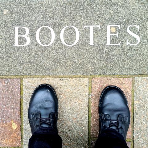 The word Bootes set into the granite paving stones on a Glasgow pavement, with the photographer's boots visible in the photo just below this word.