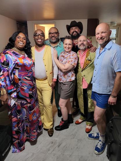 A group of seven smiling people of all shapes, sizes, colors, and genders pose in a literal rainbow of colorful clothing.