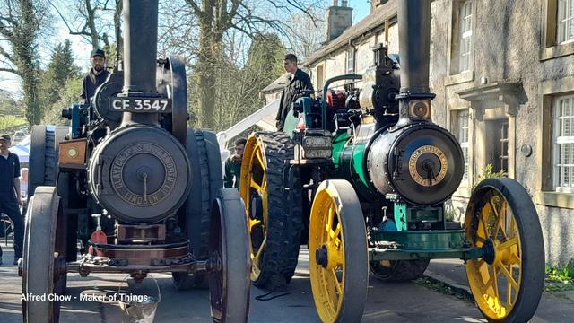 Two Clayton And Shuttleworth Ltd. steam traction engines parked side by side outside an old stone built village pub (on the right). The sky is clear blue behind the still mostly bare tree tops.
The engine on the left is seen head on, the front of the boiler framed by the smaller front, and larger rear, wheels. The registration number CF3547 is displayed at the base of the tall funnel.
The engine on the right is seen slightly from a 3/4 view so the boiler cladding frame can be seen painted in a bright green, while the wheels are painted in a bright yellow.