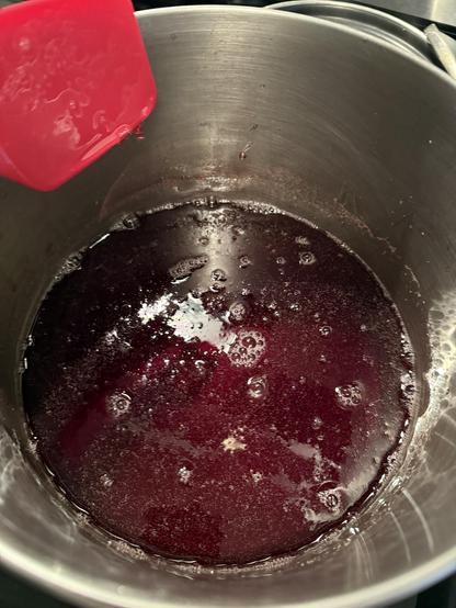 This is what the violet jelly looked like before it boiled. It's sitting in a stainless pan and it is a dark reddish purple color. There is a spatula being held up out of the jelly to the left of the frame.