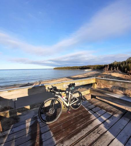 Wooden deck lookout to a sandy beach shoreline and calm lake. There is a bicycle leaning against the deck rail. The sky is blue with wispy clouds, and to the right of the lookout,  a green treed shoreline stretches out into the lake.
